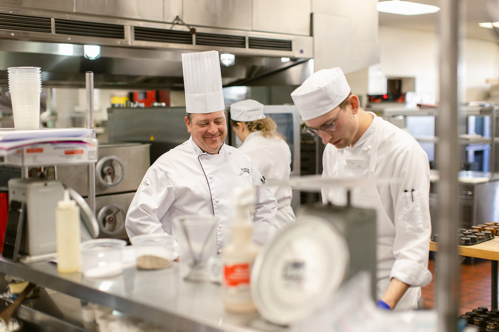 Two people in kitchen wearing chef jackets and hats.