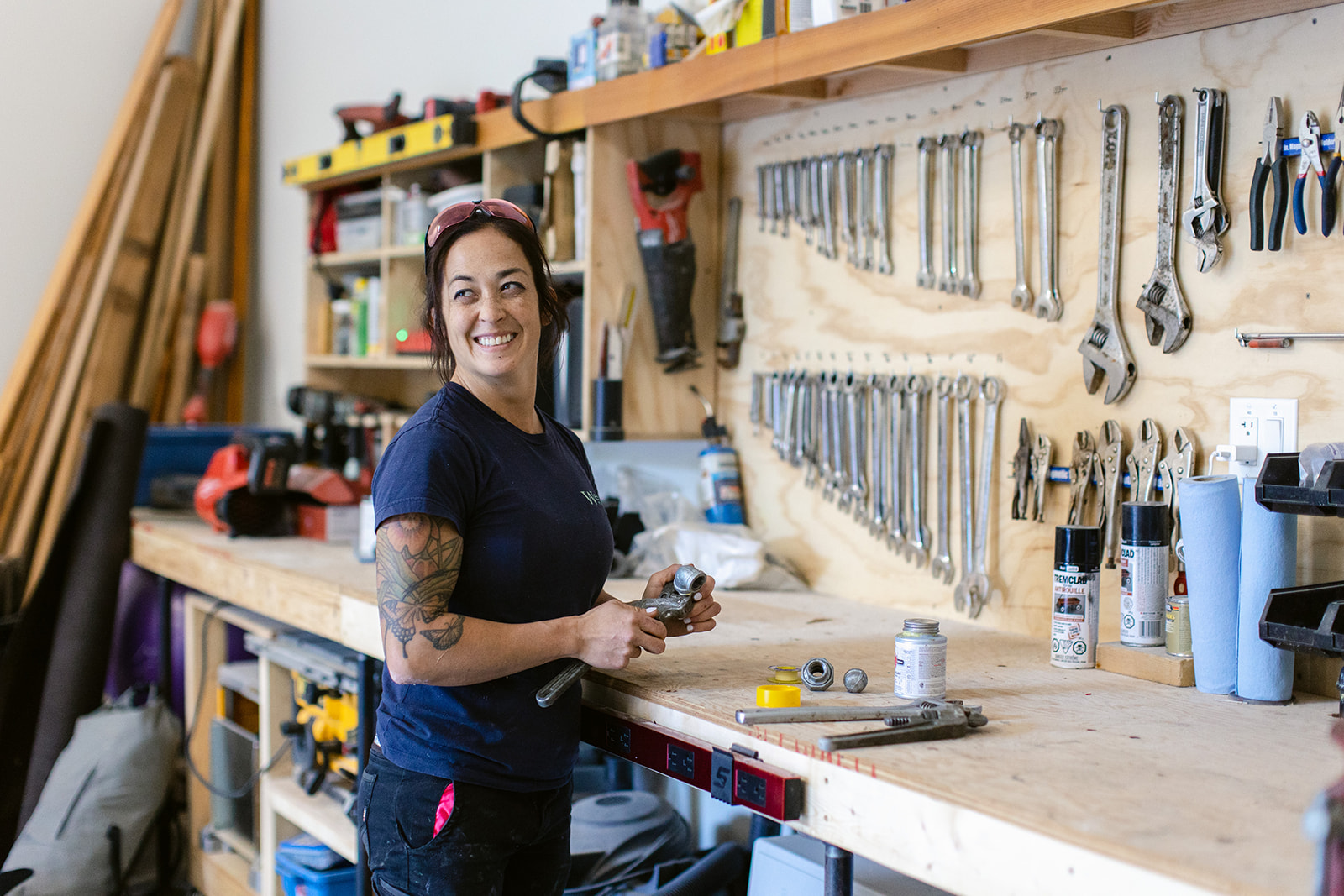 A female tradesperson stands smiling in a shop in front of a wall of tools. 
