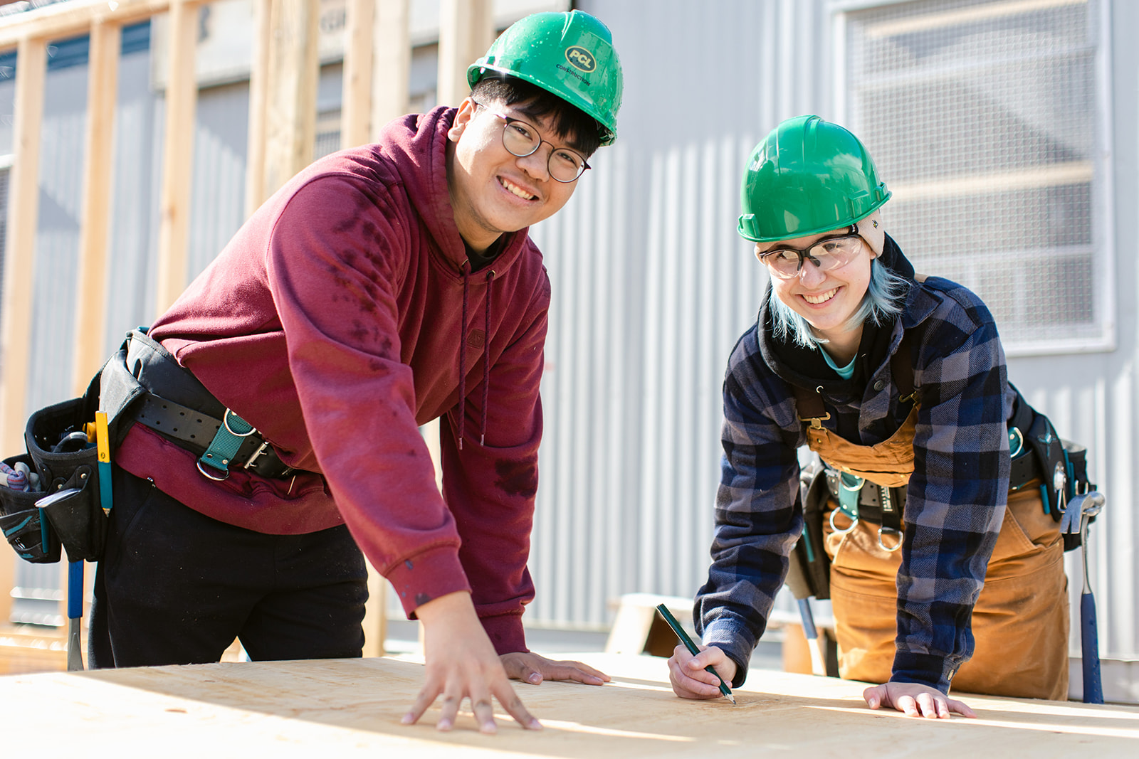 Two youth carpenters wearing green helmets smiling, writing on wooden board on site.