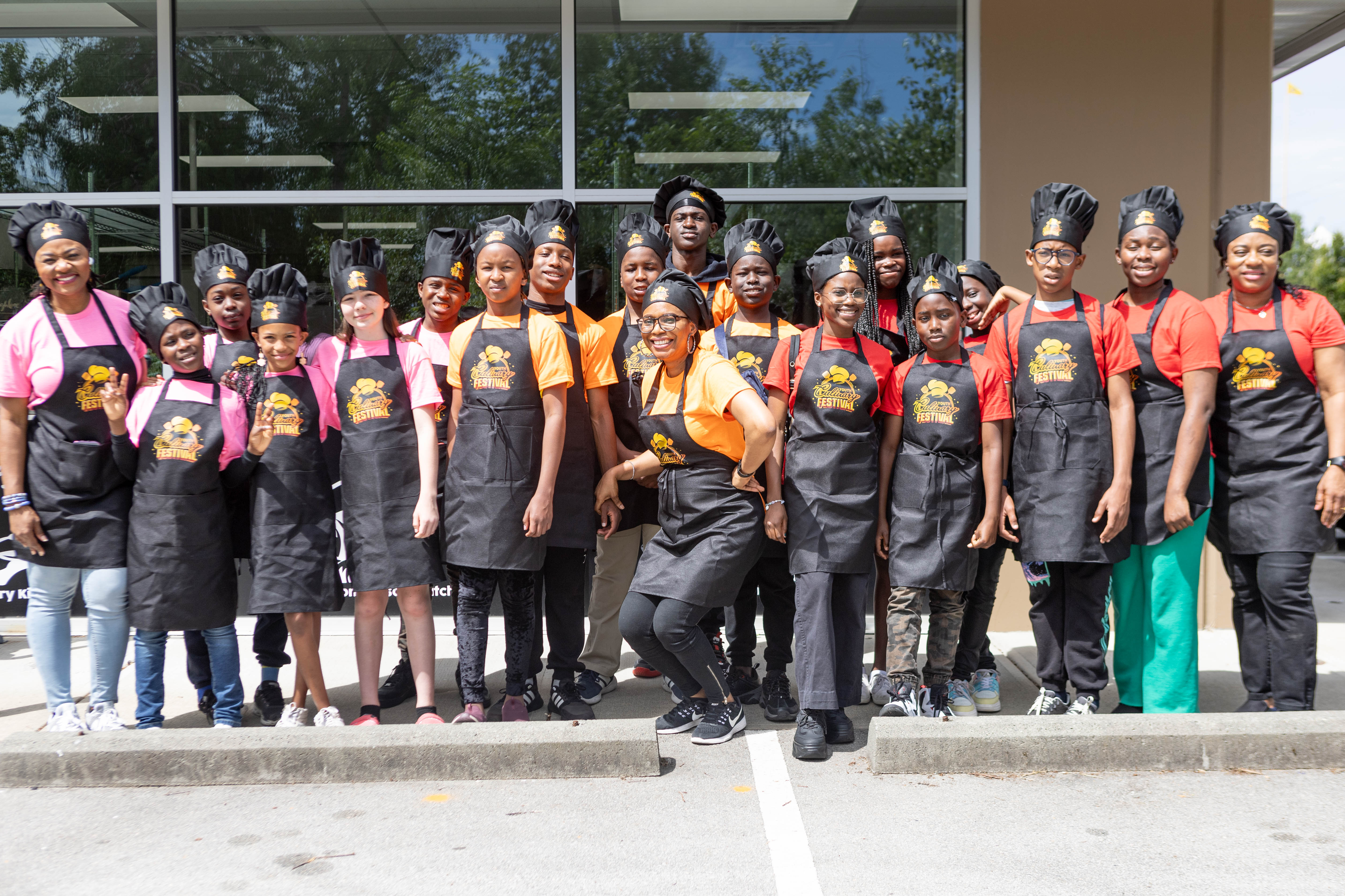 Group of people posing for photo broken into three teams, pink, orange and red wearing aprons and chef hats.