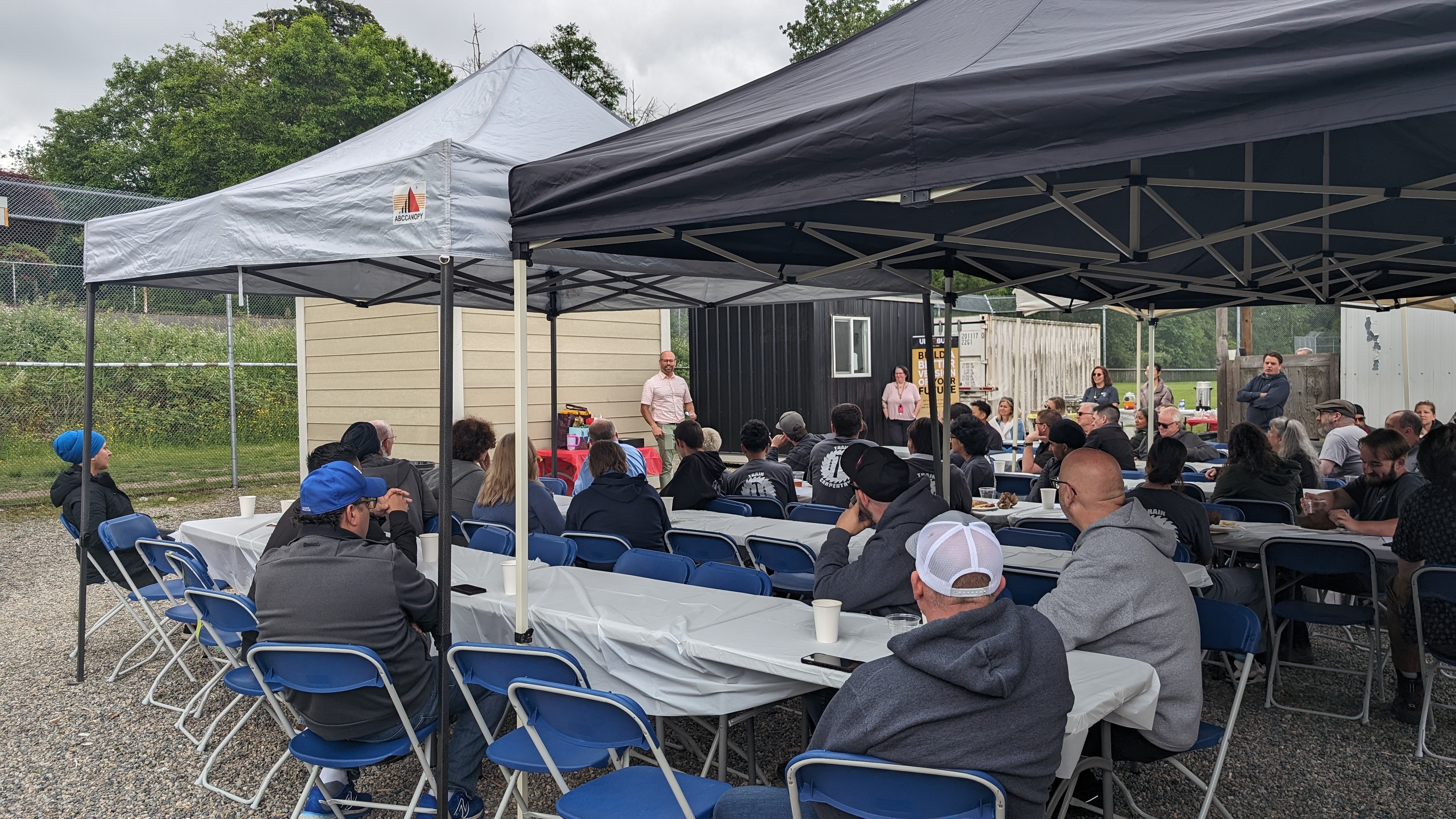 Group of people all sitting on rows of tables outside under an event tent. They are all looking away from the camera to the speaker standing in front of one of the tiny houses that were built.