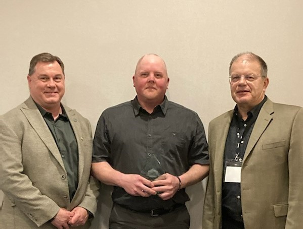 Three people standing and smiling. Middle person is holding an award