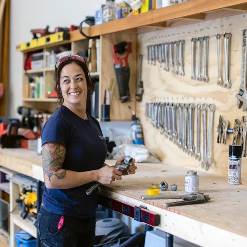A female tradesperson stands smiling in a shop in front of a wall of tools. 