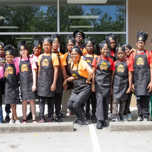 Group of people posing for photo broken into three teams, pink, orange and red wearing aprons and chef hats.